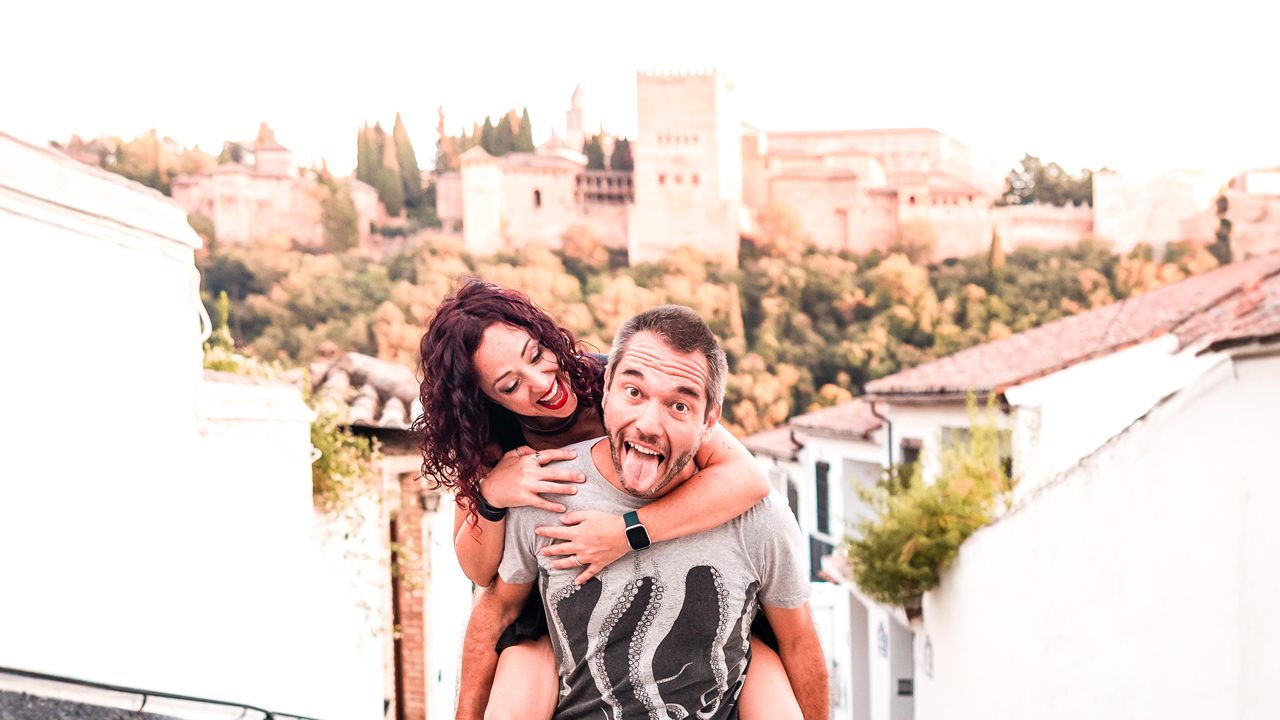UnViaggioPerDue: Daniele e Flavia a Granada, entrambi sorridenti, con gli storici edifici bianchi e l'Alhambra sullo sfondo.