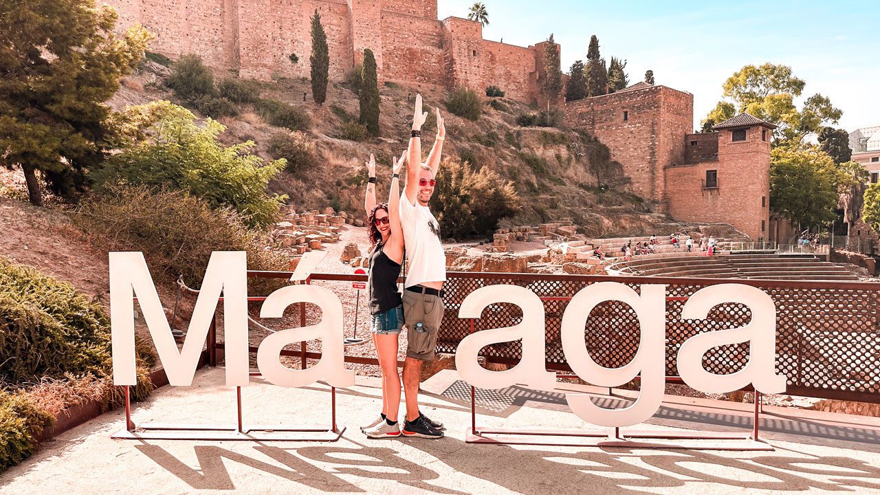 UnViaggioPerDue: Daniele e Flavia dietro un grande cartello "Malaga", con le mura storiche dell'Alcazaba sullo sfondo in una giornata di sole.
