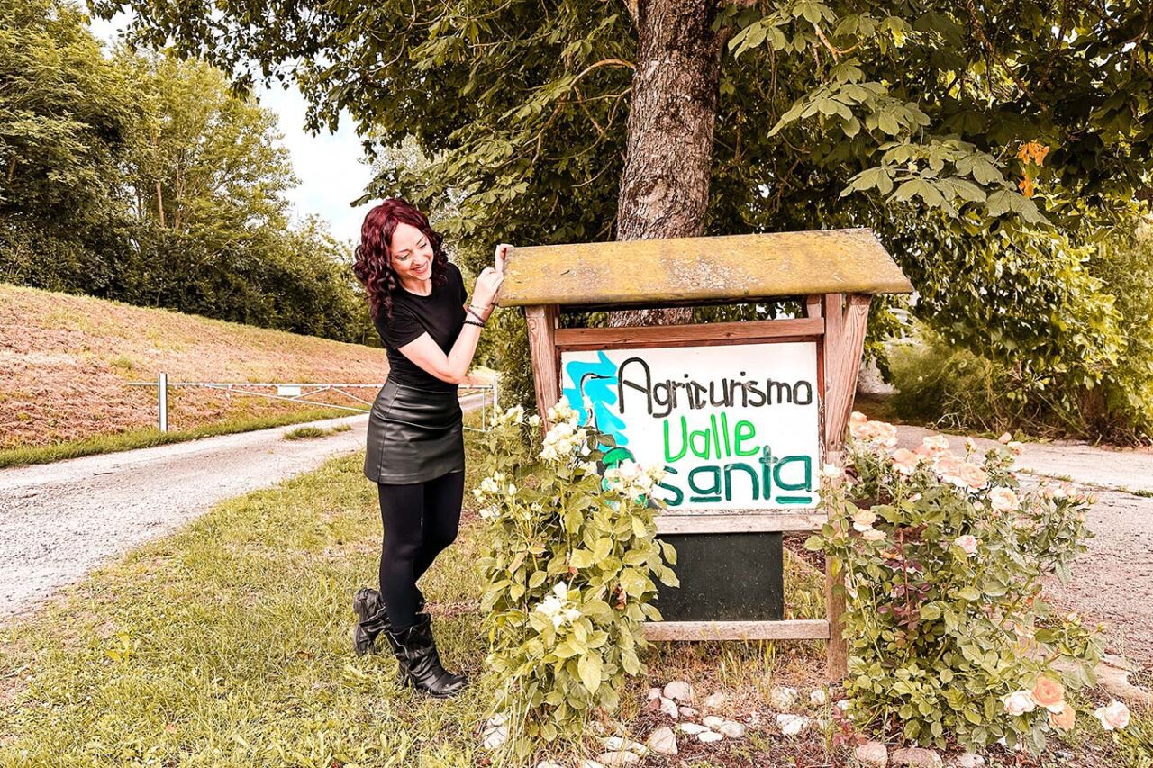 UnViaggioPerDue: Flavia accanto a un cartello in legno con la scritta "Agriturismo Valle Santa" davanti a un albero su un sentiero rurale immerso nel verde, catturando il sereno fascino rustico della provincia di Ferrara.