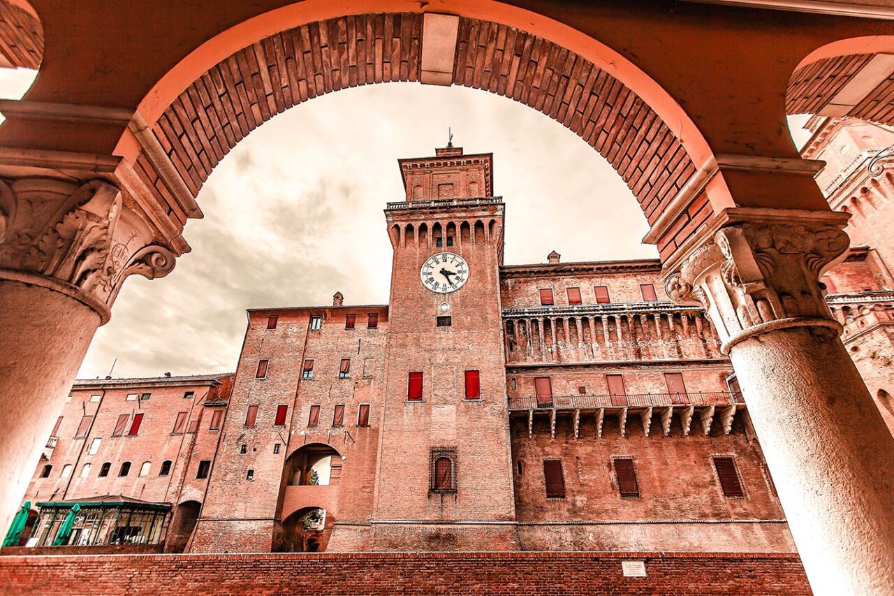 UnViaggioPerDue: Castello medievale in mattoni con una torre dell'orologio, visto attraverso un colonnato ad arco caratterizzato da intricati dettagli sui suoi pilastri sotto un cielo nuvoloso a Ferrara.