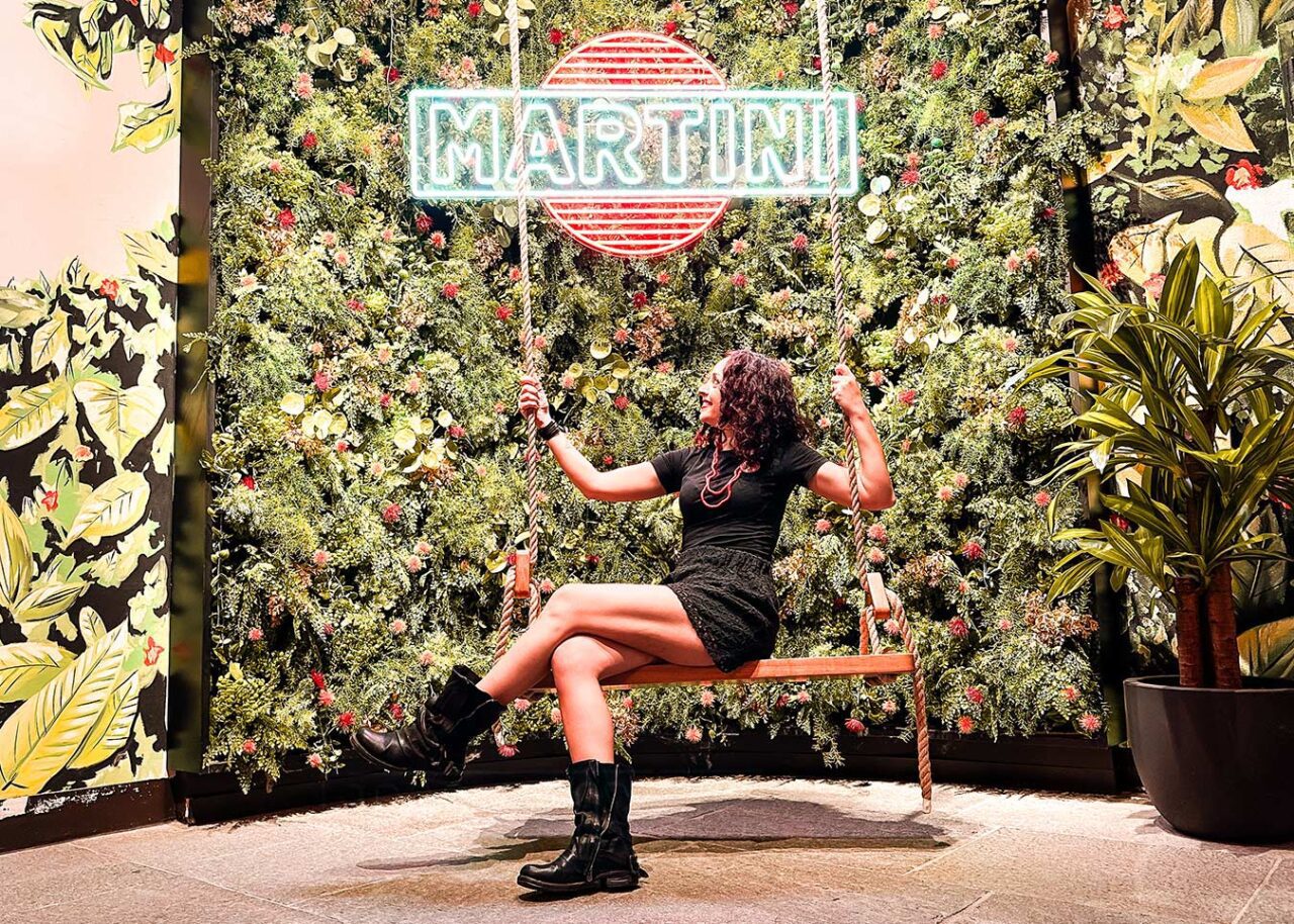 UnViaggioPerDue: Flavia seduta su un'altalena davanti a un muro verde adornato con piante decorative e un'insegna al neon "Martini", catturando l'essenza di Casa Martini a Torino.