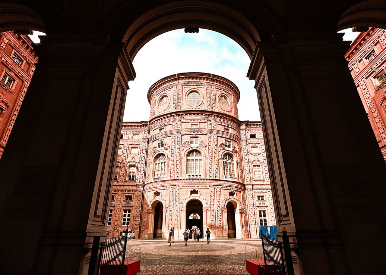 UnViaggioPerDue: Vista attraverso un arco dello storico e ornato Palazzo Carignano a Torino, con grandi finestre e dettagli intricati; alcune persone camminano nel cortile.