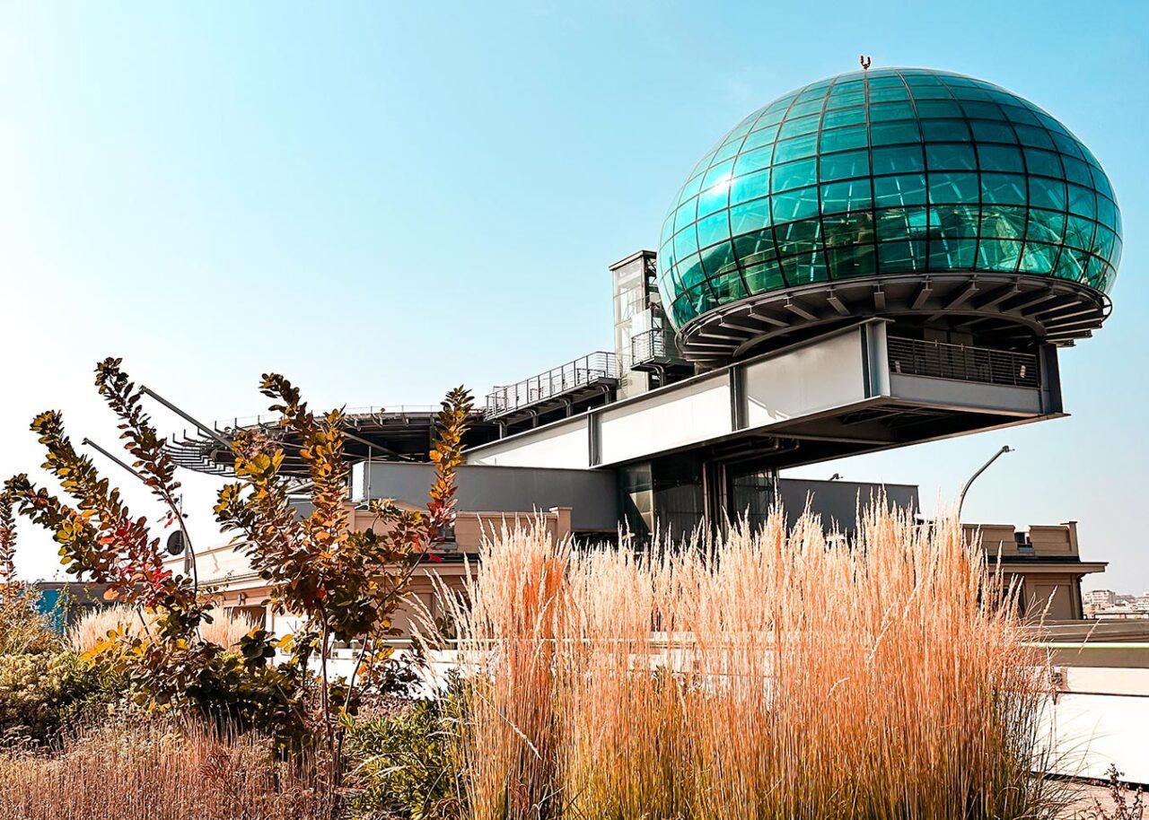 UnViaggioPerDue: Un edificio dal design futuristico è caratterizzato da una grande cupola di vetro blu-verdastro e da una struttura metallica portante. Questo edificio dal design innovativo si trova sulla Pista 500 di Torino, circondato da erba e piante sotto un cielo azzurro e limpido.