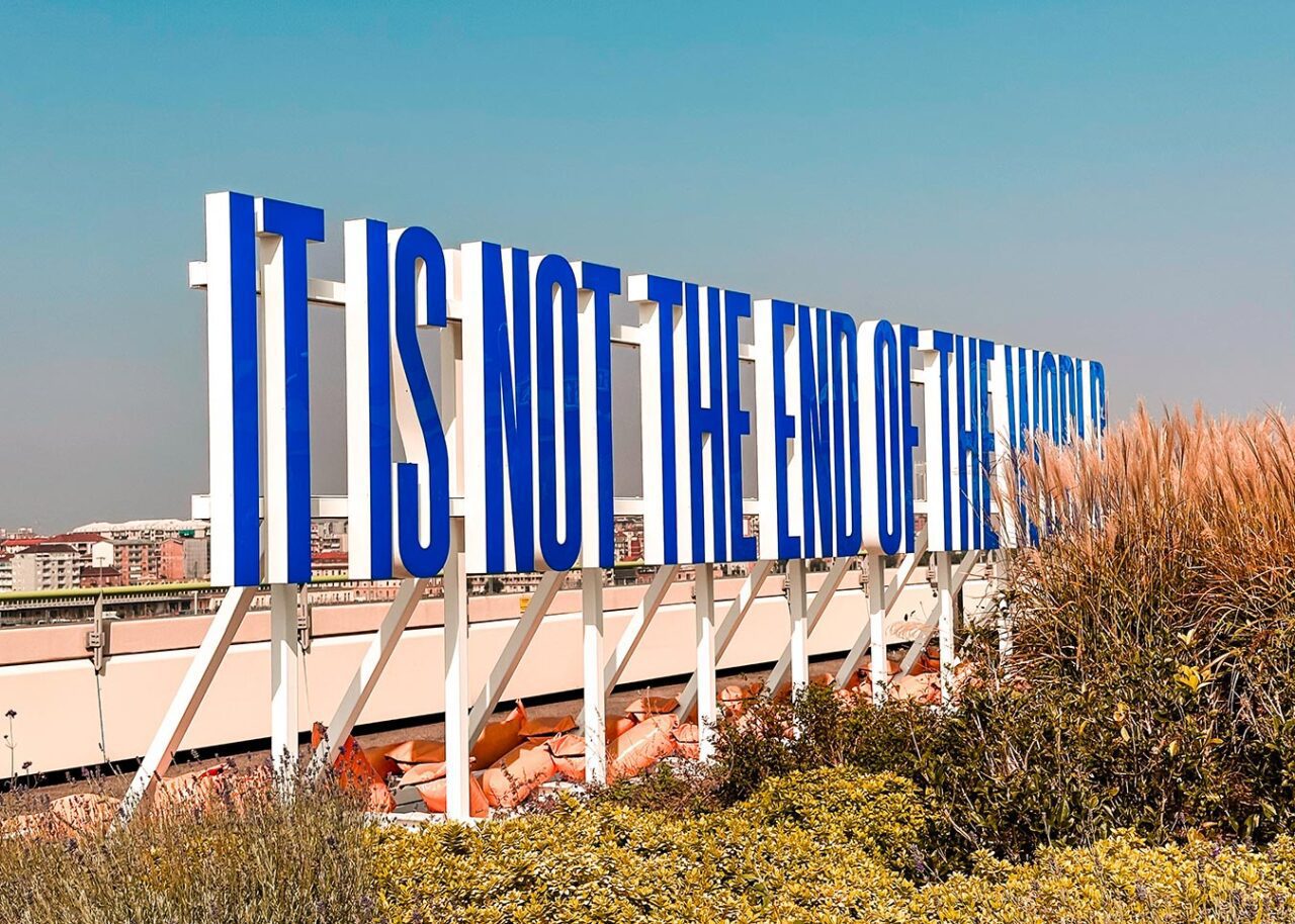 UnViaggioPerDue: Installazione lungo la Pista 500 di Torino con un grande cartello blu e bianco recita "NON È LA FINE DEL MONDO" contro un cielo limpido, con edifici e fogliame sullo sfondo.