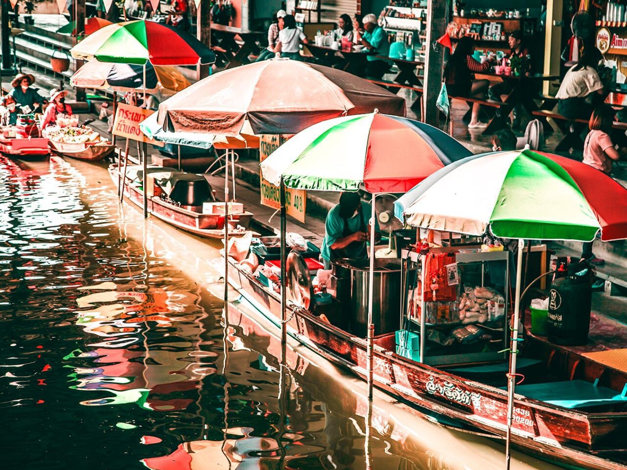 UnViaggioPerDue: Diversi venditori vendono merci da barche con ombrelli colorati su un vivace fiume al mercato galleggiante di Damnoen. Le persone fanno acquisti e interagiscono nelle vicinanze.