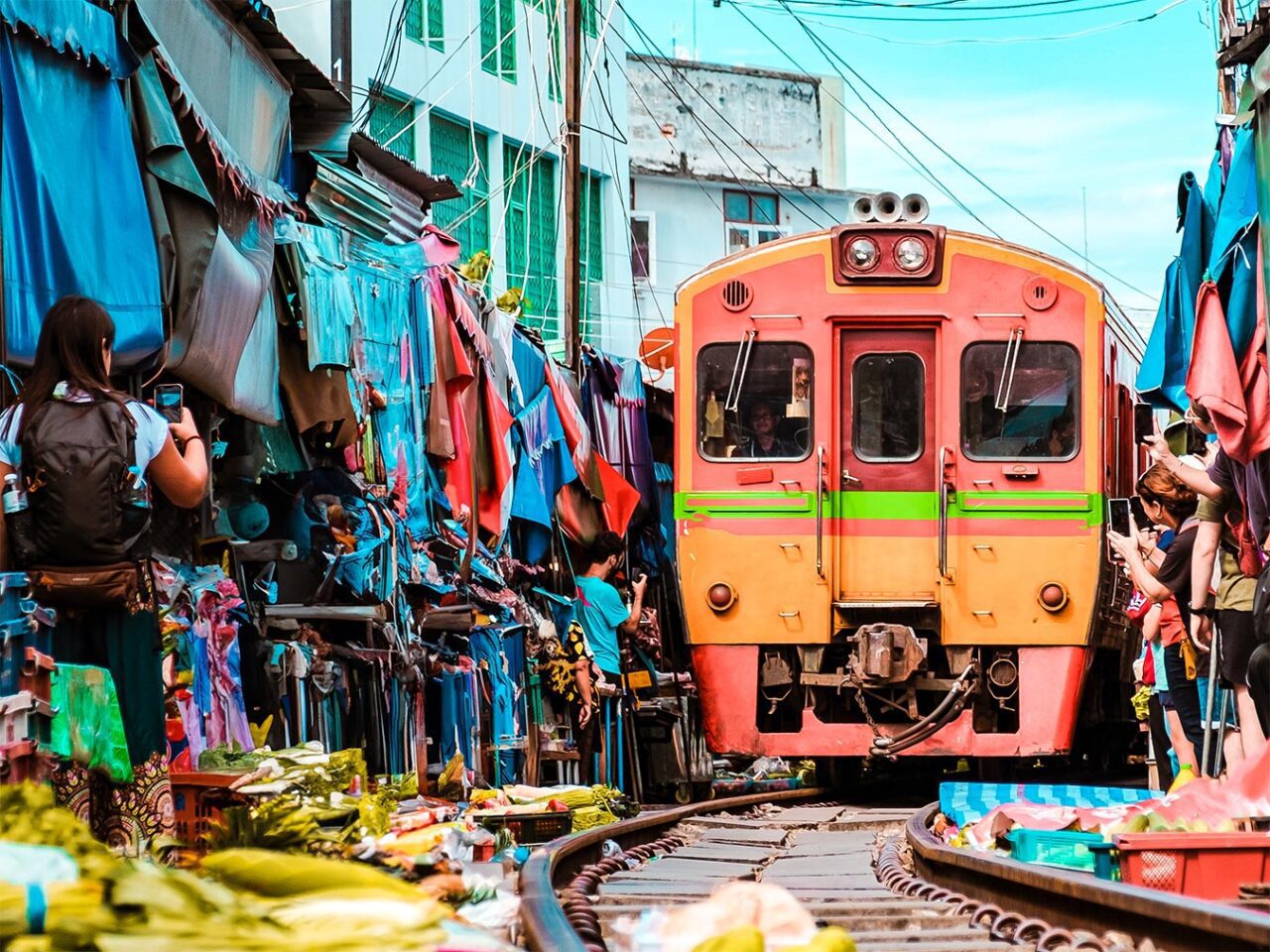 UnViaggioPerDue: Un treno attraversa la stretta strada del mercato ferroviario di Maeklong, fiancheggiata da bancarelle e tessuti colorati, con persone che scattano foto e vendono merci nelle vicinanze.