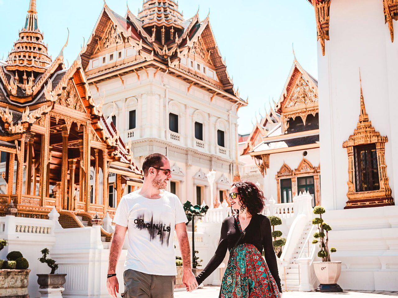 UnViaggioPerDue: Daniele e Flavia, tenendosi per mano, passeggiano attraverso un complesso di templi con edifici decorati in oro all'interno del Palazzo Reale di Bangkok. Sullo sfondo in una giornata soleggiata.