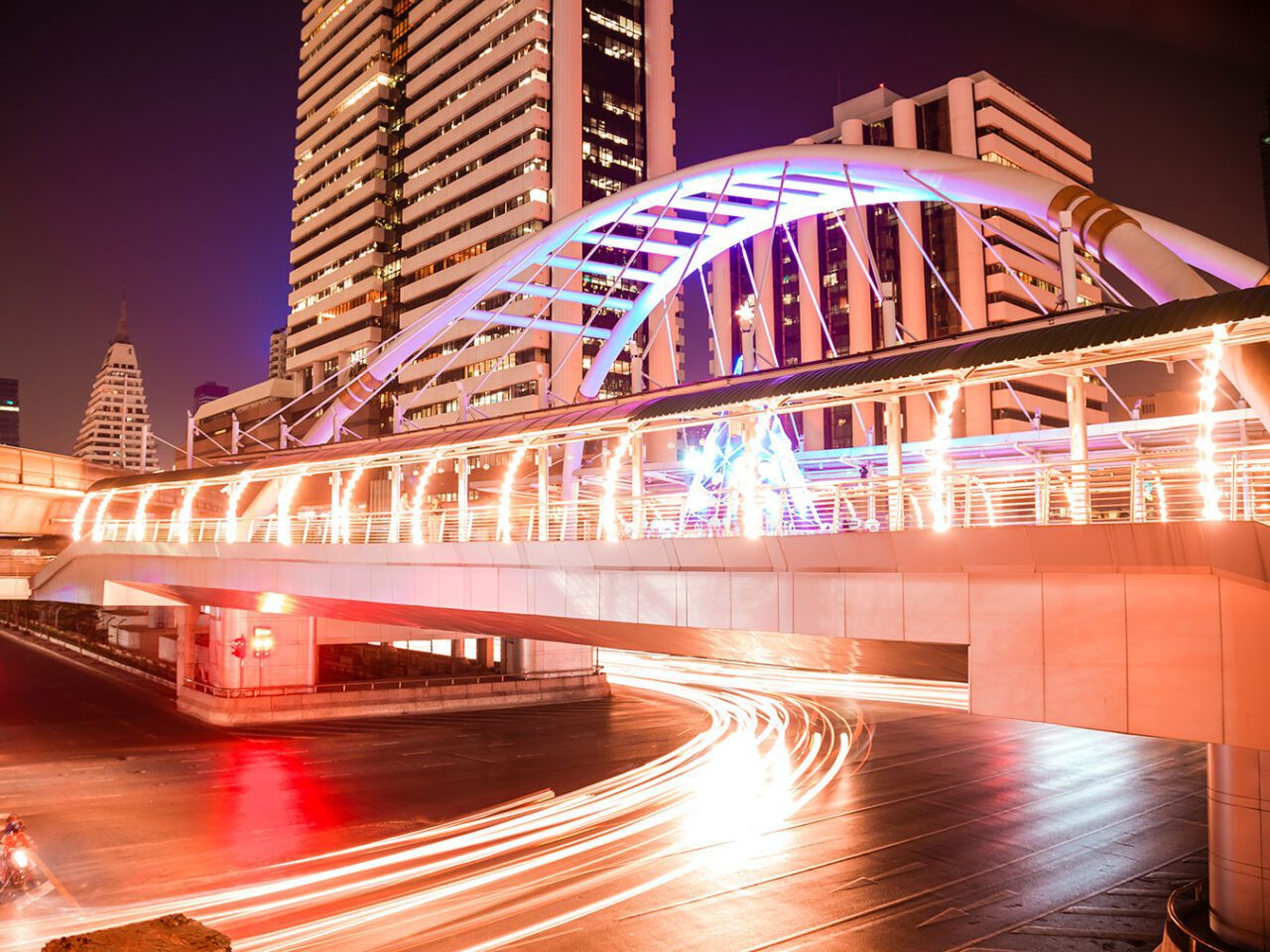 UnViaggioPerDue: Moderno ponte pedonale illuminato dal design curvo su Sliom Road, una strada trafficata di Bangkok, strisce di luci dei veicoli catturate con una lunga esposizione e alti edifici sullo sfondo di notte.