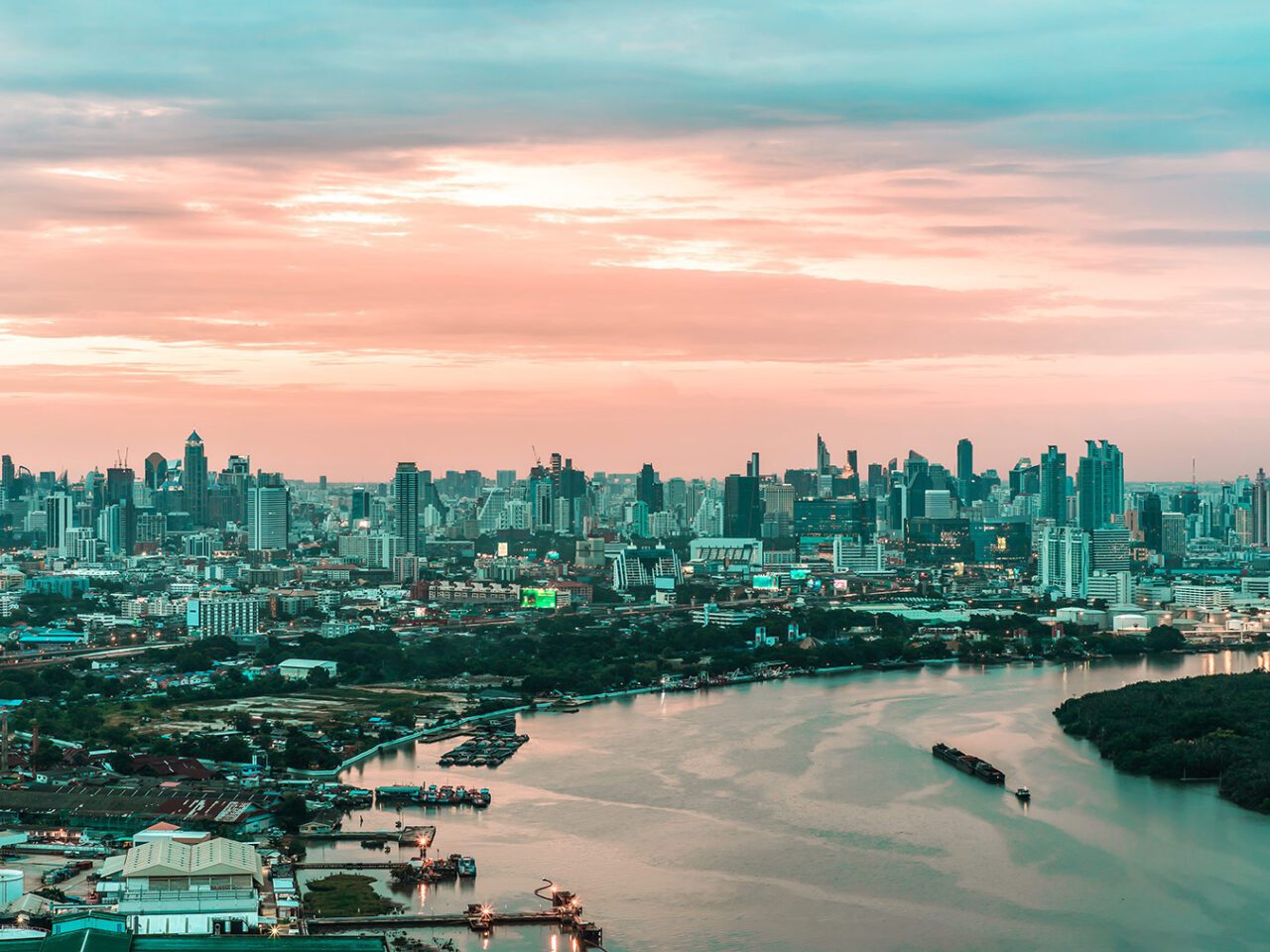 UnViaggioPerDue: Uno skyline di Bangkok al tramonto presenta molti edifici alti e un fiume in primo piano. Il cielo è carico di nuvole, illuminato dai colori pastello del tramonto.