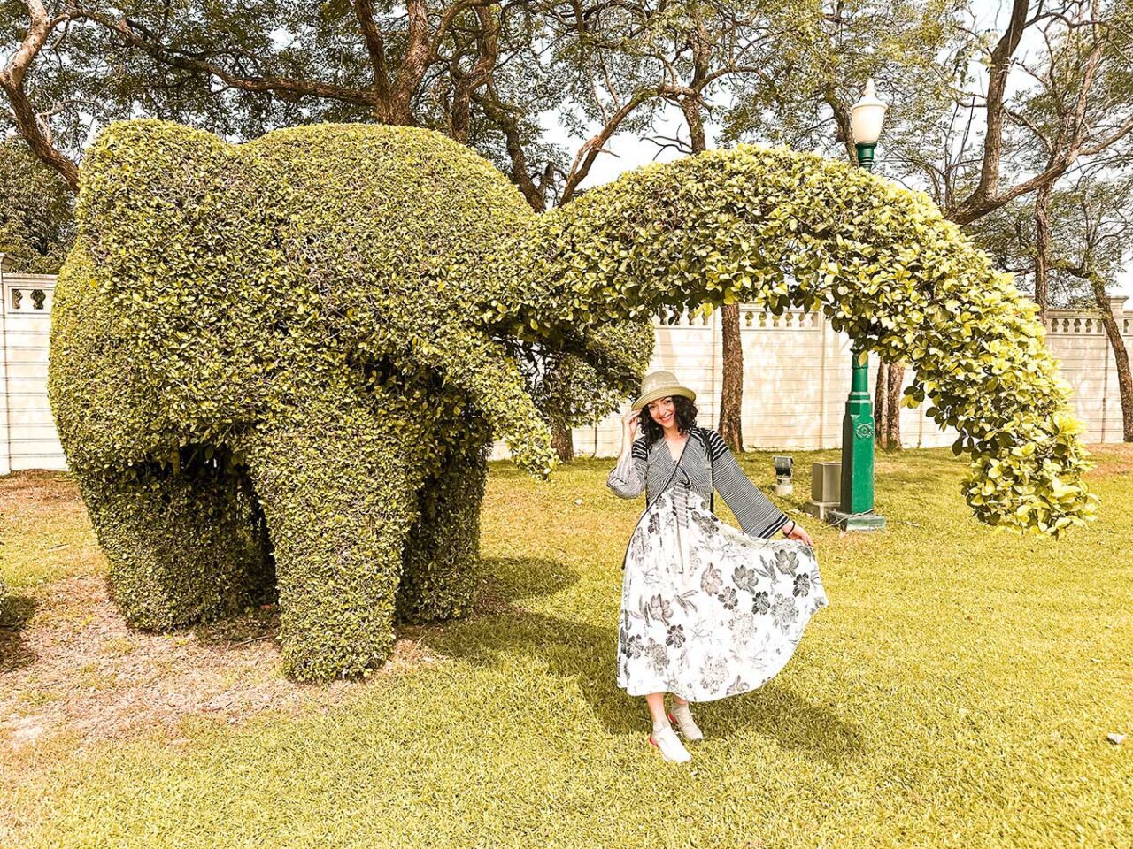 UnViaggioPerDue: Flavia con un abito floreale bianco e un cappello da sole posa accanto a una grande arte topiaria a forma di elefante, all'interno di un lussureggiante giardino della Thailandia.