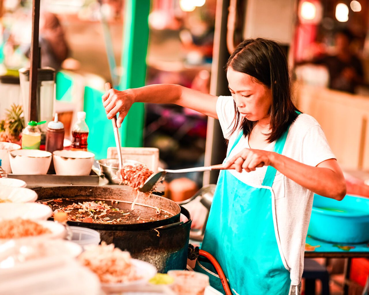 UnViaggioPerDue: Una persona con un grembiule blu sta cucinando cibo di strada thailandese in un grande mercato all'aperto, mescolando una pentola su un fornello.