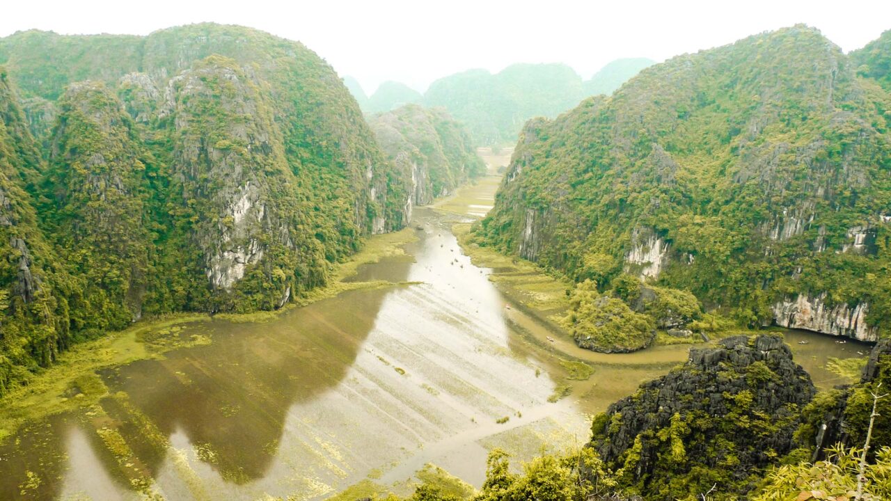 UnViaggioPerDue: vista aerea del Fiume Tam Coc in Vietnam, che si snoda tra rigogliose e ripide colline verdi e scogliere rocciose, con alcune zone di acqua bassa e vegetazione lungo le rive del fiume.