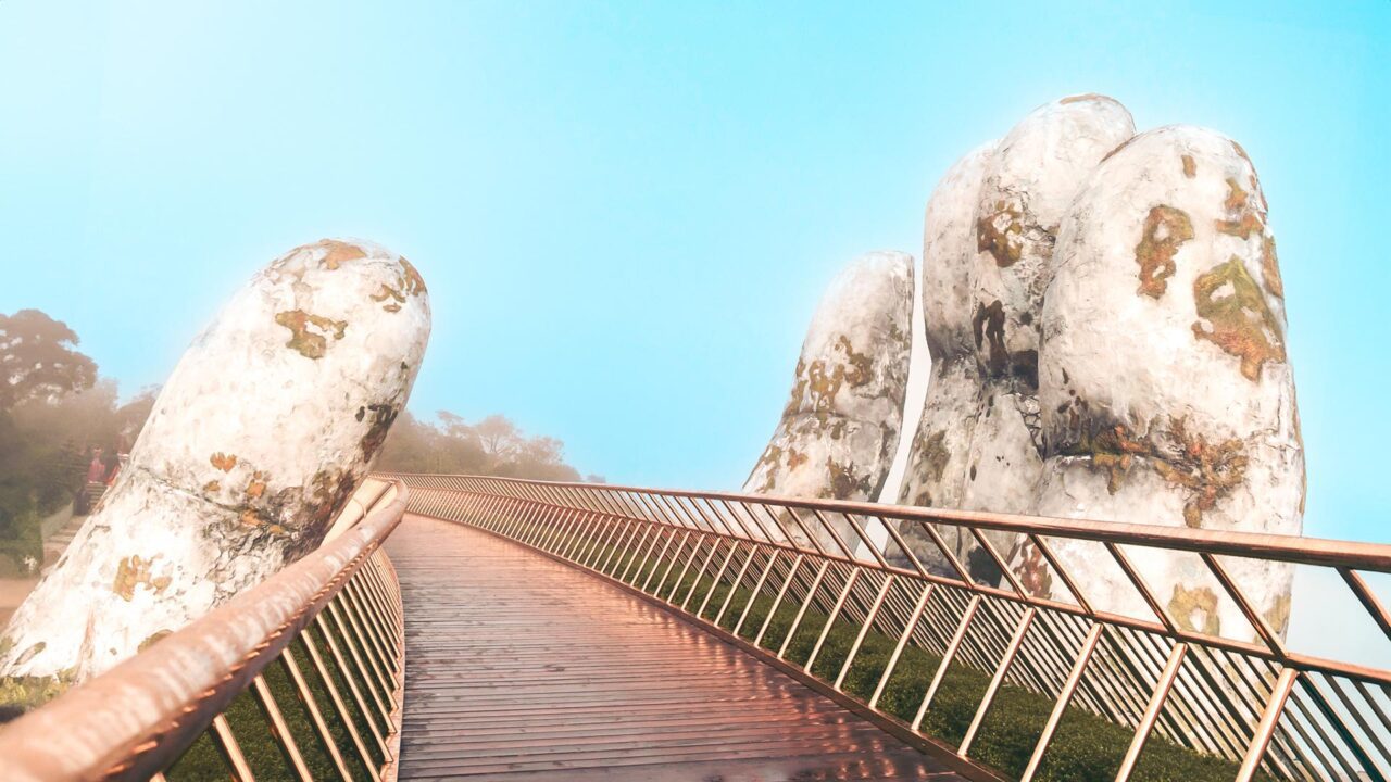 UnViaggioPerDue: ponte pedonale, noto come Golden Hand Bridge in Vietnam, è sorretto da grandi sculture di mani in pietra che si stendono dal basso. Il cielo è di un azzurro limpido.