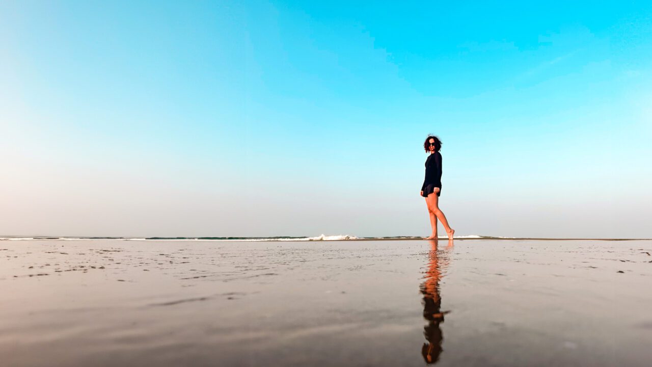 UnViaggioPerDue: Flavia vestita di scuro in piedi su una spiaggia ampia e bagnata, con un cielo azzurro e limpido sopra, sulle tranquille rive di Hoi An, Vietnam.