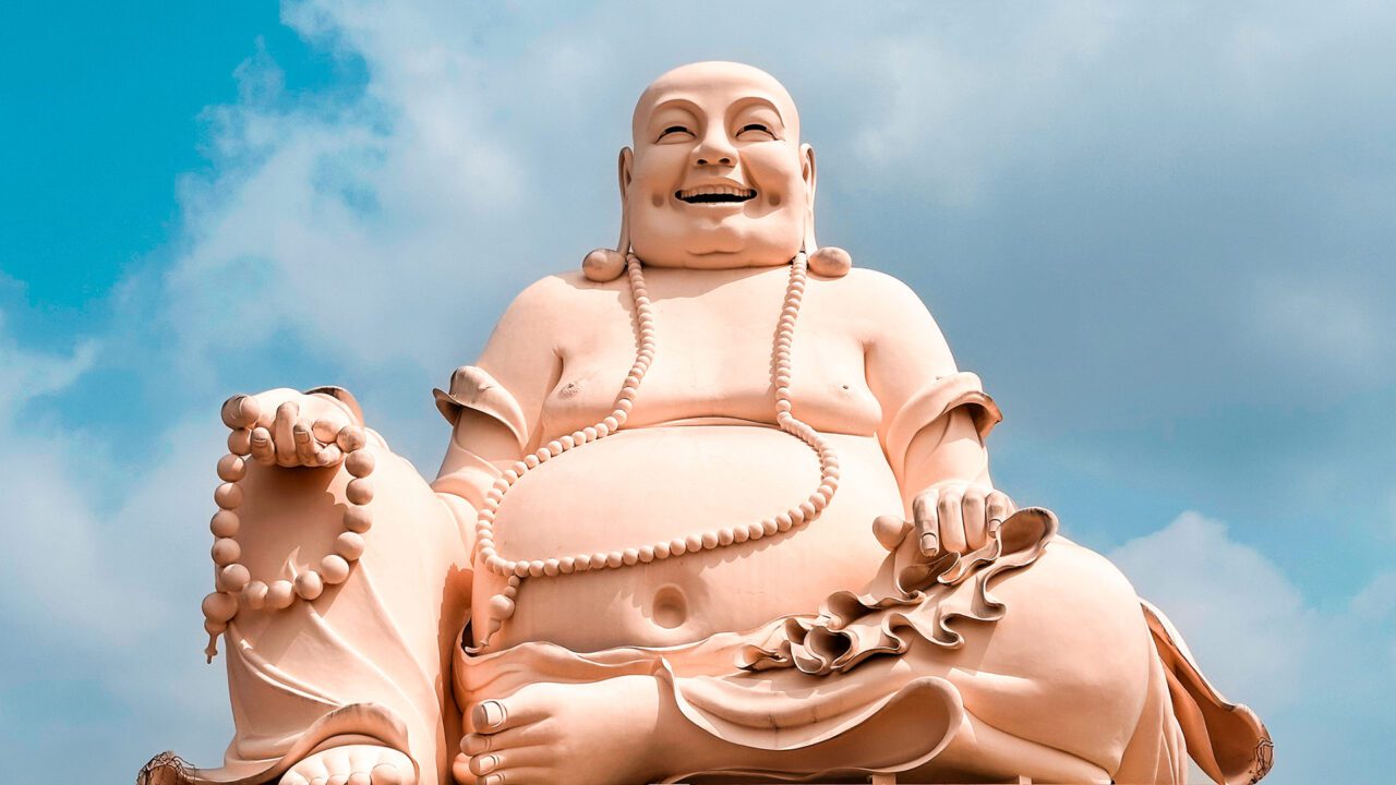 UnViaggioPerDue: una grande statua di un Buddha sorridente con una collana di perline è seduta comodamente sullo sfondo di un cielo azzurro parzialmente nuvoloso, Pagoda di Vinh Trang in Vietnam.