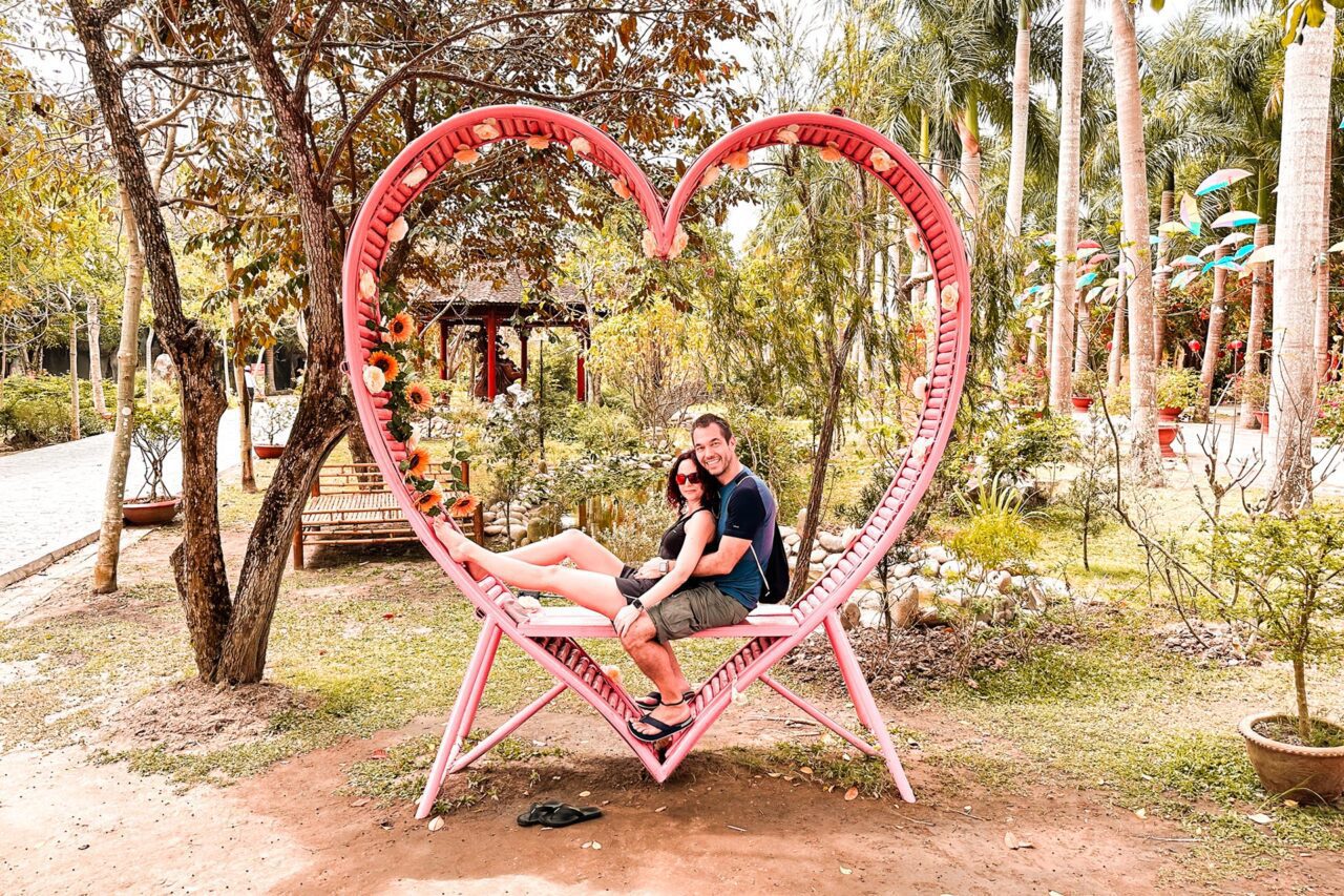 UnViaggioPerDue: Daniele e Flavia sono seduti su una panchina rosa a forma di cuore in un parco, circondati da una vegetazione lussureggiante che ricorda la bellezza naturale del Vietnam.